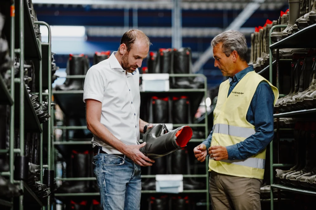Twee personen in een opslagruimte met rekken vol zwarte werkschoenen bespreken voorraadbeheer en de voordelen van inkoop uitbesteden